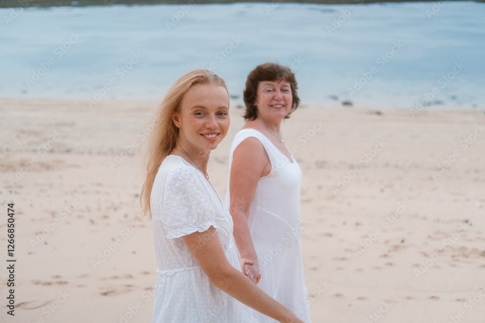 A senior mother and her adult redhead daughter in white dresses holding hands are walking along the beach by the ocean. They are talking to each other and laughing. Family vacation. Mothers Day. A senior mother and her adult redhead daughter in white dresses holding hands are walking along the beach by the ocean. They are talking to each other and laughing. Family vacation. Mothers Day.