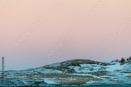 A bright orange glow colours the golden hour sky that serves as a dramatic background for the rugged mountains and snow-covered beaches of arctic norway, near the town of Bodo. photo