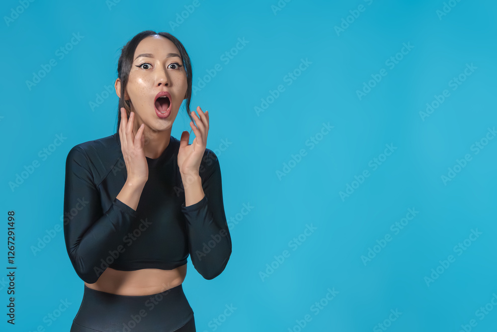 Slender r Asian girl in sportswear expresses surprise or shock. Her eyes are wide open, her mouth is open, and her hands are raised to her face in an emotional gesture. Isolated blue background. Slender r Asian girl in sportswear expresses surprise or shock. Her eyes are wide open, her mouth is open, and her hands are raised to her face in an emotional gesture. Isolated blue background.