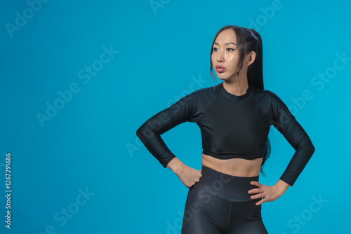 Slender beautiful Asian girl in sportswear has taken a confident pose, putting her hands on her waist, and is looking away with slight thoughtfulness. Confidence and concentration