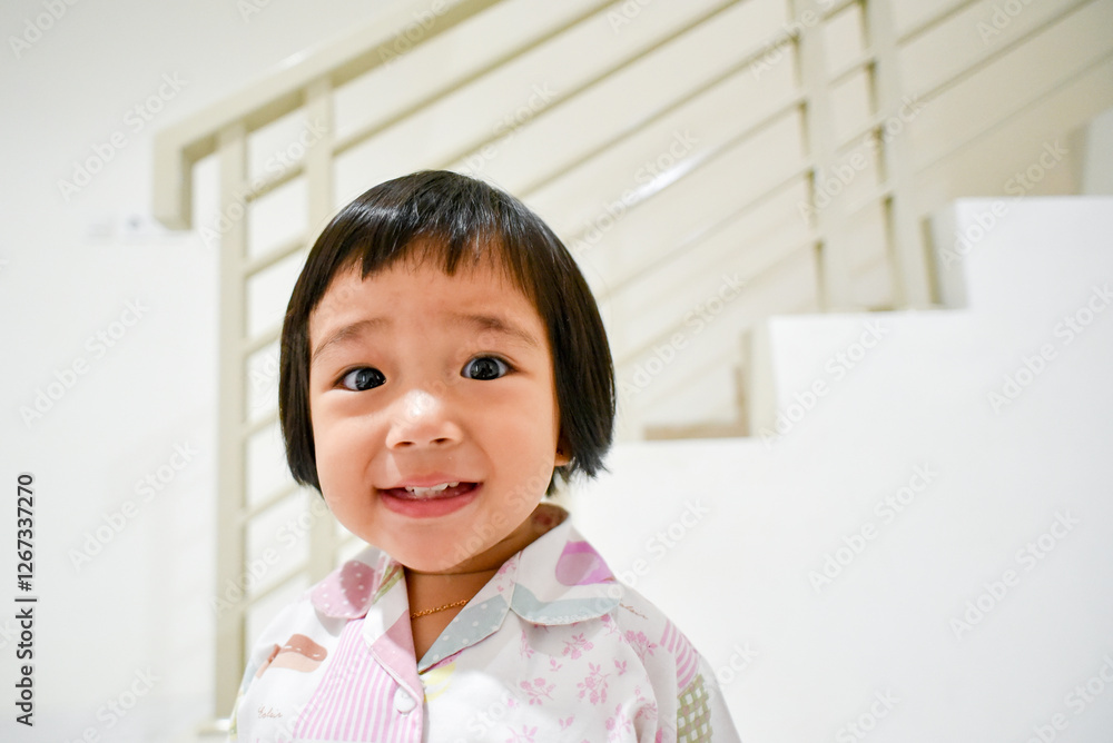 A close-up portrait of a joyful Asian toddler with short black hair, wearing pastel-colored pajamas, smiling brightly against a modern white staircase background A close-up portrait of a joyful Asian toddler with short black hair, wearing pastel-colored pajamas, smiling brightly against a modern white staircase background