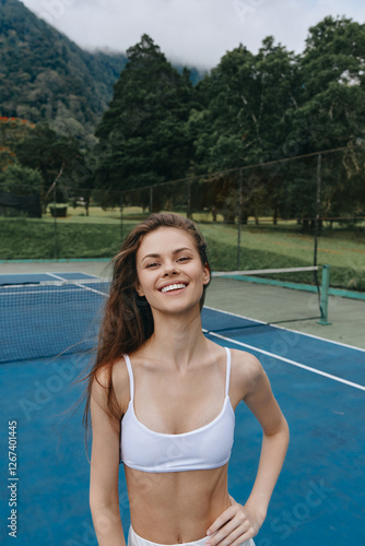Joyful young woman in a white sports bra enjoying a tennis match on a vibrant outdoor court with lush green trees in the background