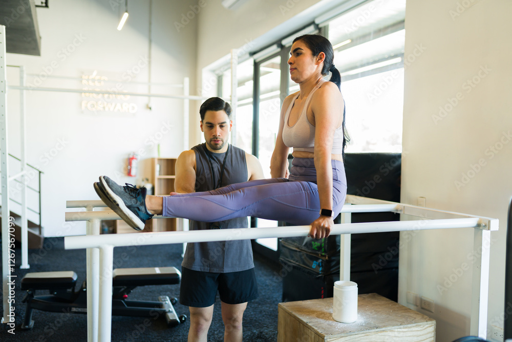 Fototapeta premium Latina fitness enthusiast performing challenging l-sit core workout with trainer guidance during strength training session at gym