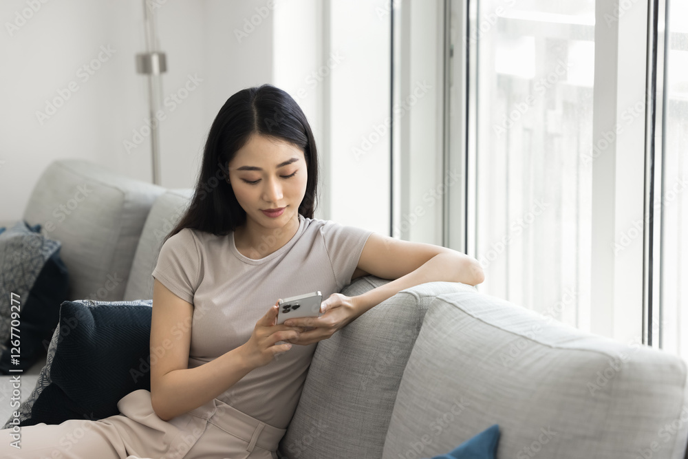 Focused Chinese woman resting on sofa with smartphone, shopping online, browsing e-shopping websites, watching videos via streaming platforms, checking emails, reply to messages through applications Focused Chinese woman resting on sofa with smartphone, shopping online, browsing e-shopping websites, watching videos via streaming platforms, checking emails, reply to messages through applications