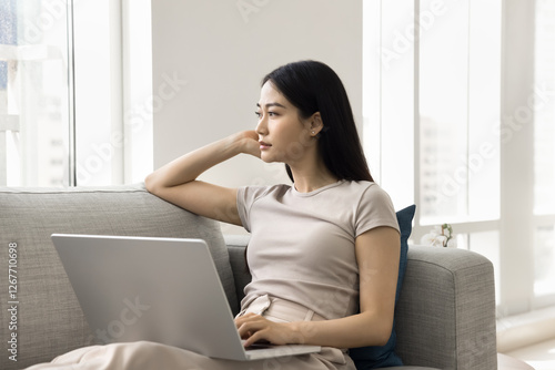 Obraz na plátně Calm woman gazing thoughtfully into distance seated on sofa with laptop, in moment of deep reflection, contemplation, planning, decision-making, search solutions, strategizing about tasks