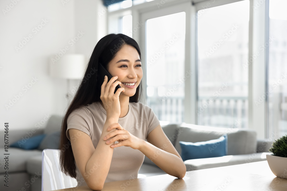 Beautiful Asian woman sitting at table in cozy living room, smile, lead pleasant conversation on smartphone, receive good news, enjoy positive communication remotely by phonecall. Modern technology Beautiful Asian woman sitting at table in cozy living room, smile, lead pleasant conversation on smartphone, receive good news, enjoy positive communication remotely by phonecall. Modern technology
