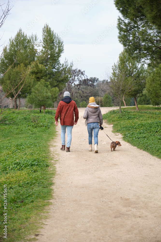 Couple walking with their dachshund dog in a park path during a cloudy day Couple walking with their dachshund dog in a park path during a cloudy day