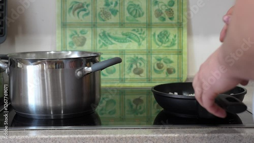 A man cooks on an induction hob, stirring chopped onions in a frying pan while a pot simmers on a nearby burner. The scene shows the process of preparing a home-cooked meal