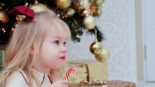 Caucasian child toddler girl in pajamas licks lollipop in front of the Christmas tree at home.