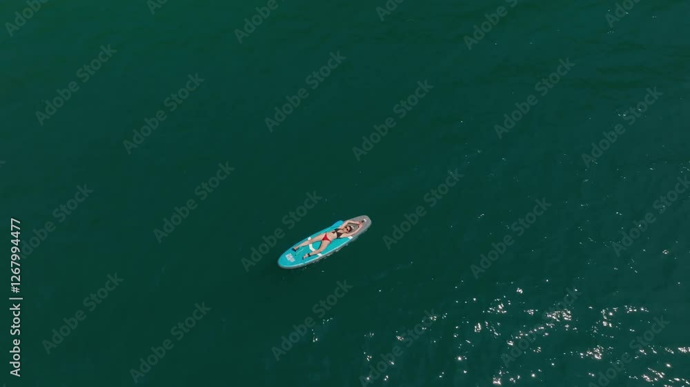 Drone view pulling away from a woman in a swimsuit lying on a SUP board, enjoying the summer in the blue waters of Montenegro Drone view pulling away from a woman in a swimsuit lying on a SUP board, enjoying the summer in the blue waters of Montenegro