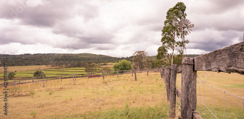 Rustic Australian farm landscape featuring a weathered wooden fence and windmill under a cloudy sky. A classic rural scene capturing the charm of the countryside.