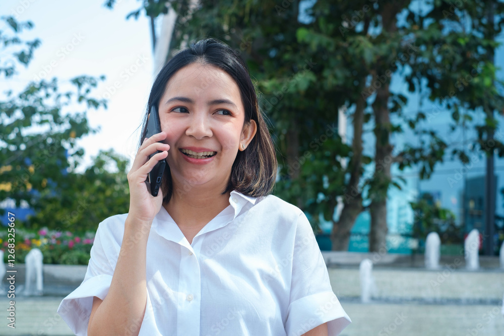 Smiling woman in white shirt having a phone conversation outdoors, surrounded by greenery and urban architecture, expressing communication and happiness. Smiling woman in white shirt having a phone conversation outdoors, surrounded by greenery and urban architecture, expressing communication and happiness.