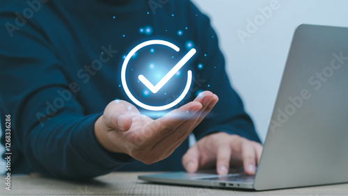 A hand showcasing a glowing check mark symbol, representing success and approval in a modern digital workspace with a laptop.