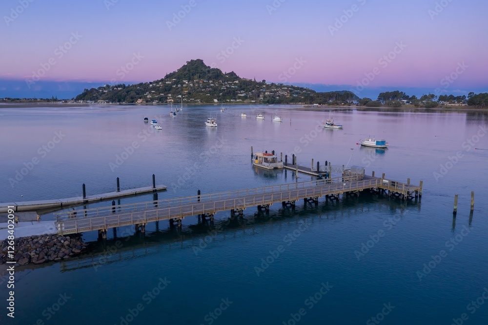 Obraz premium Wooden pier extends into calm water, boats moored nearby, tranquil coastal scene at dusk. Tairua, Coromandel Peninsula, New Zealand