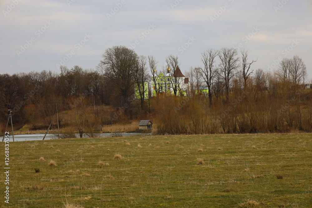 Fototapeta premium Beautiful landscape with an old church on a hill covered with trees near a picturesque pond. Cloudy weather.