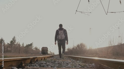 A young man with a hook and backpack walks along railroad tracks at sunset and looks away from the camera.