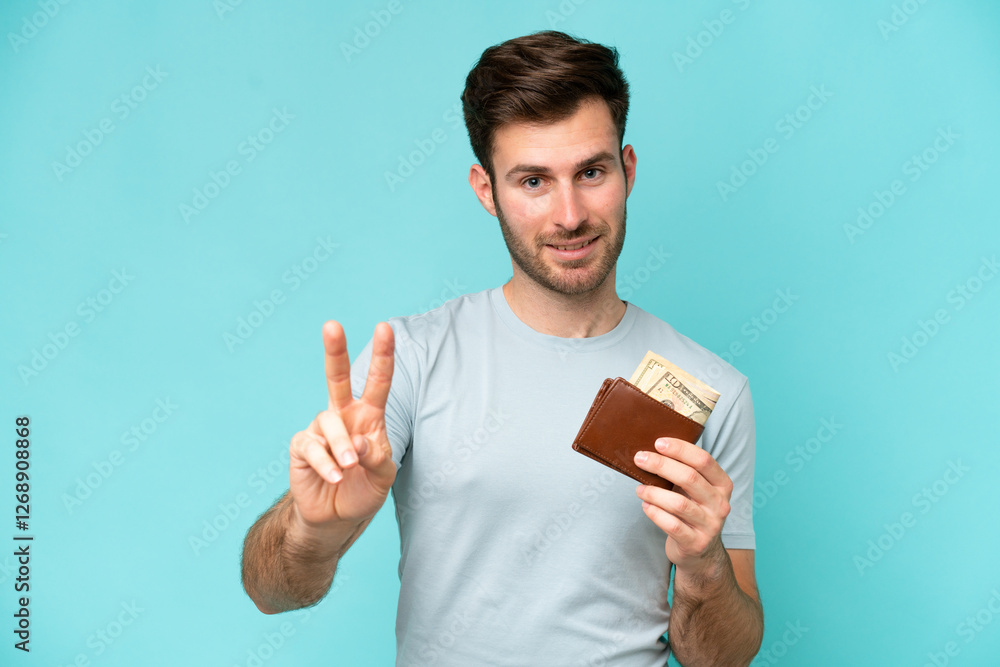 Young caucasian man holding a wallet isolated on blue background smiling and showing victory sign Young caucasian man holding a wallet isolated on blue background smiling and showing victory sign