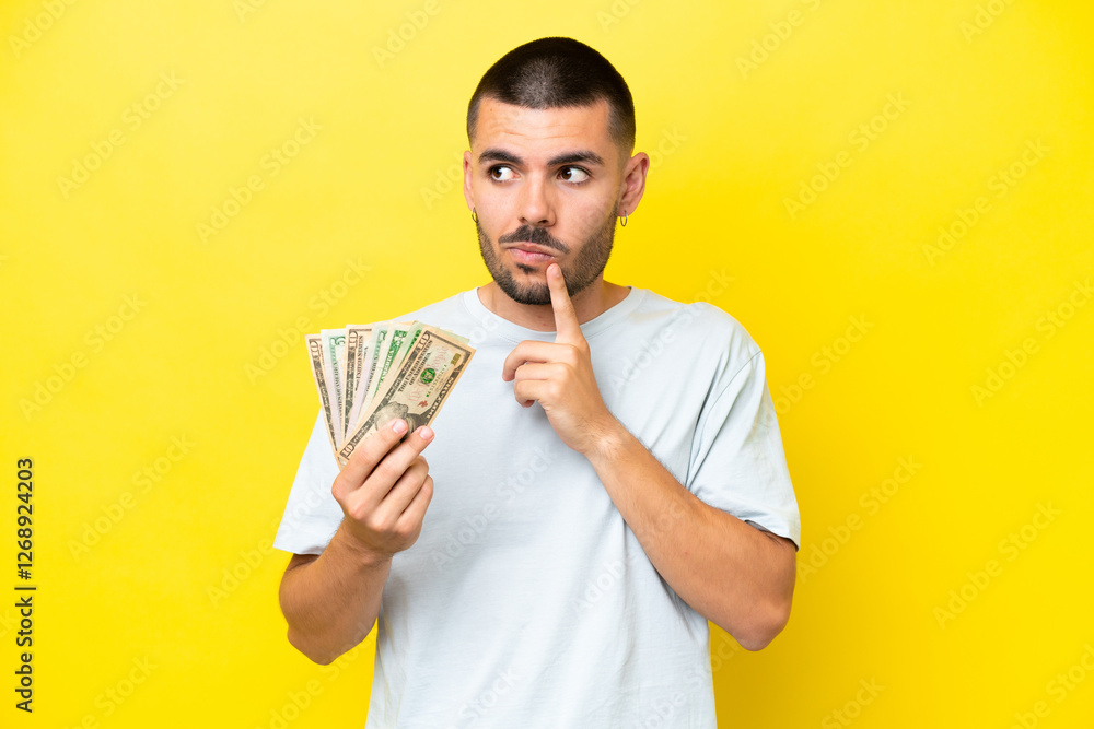 Young caucasian man taking a lot of money isolated on yellow background having doubts and thinking Young caucasian man taking a lot of money isolated on yellow background having doubts and thinking