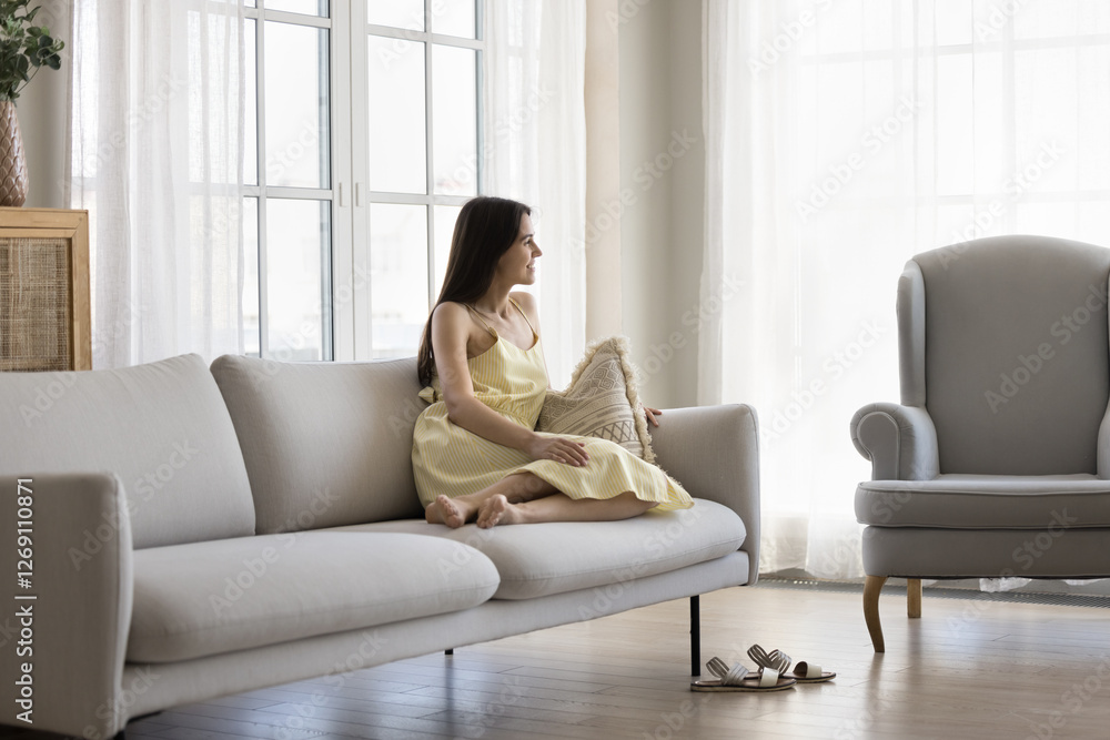Enjoying wellbeing. Happy smiling pretty young girl in summer dress relax sitting on couch in light luxury living room interior look away dreaming of future satisfied with elegant comfort of own house
