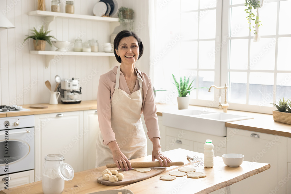 Expert in domestic culinary. Portrait happy smiling aged lady in apron looking at camera taking break in preparing home cooked meal flattening dough for baking buns in contemporary kitchen. Copy space Expert in domestic culinary. Portrait happy smiling aged lady in apron looking at camera taking break in preparing home cooked meal flattening dough for baking buns in contemporary kitchen. Copy space