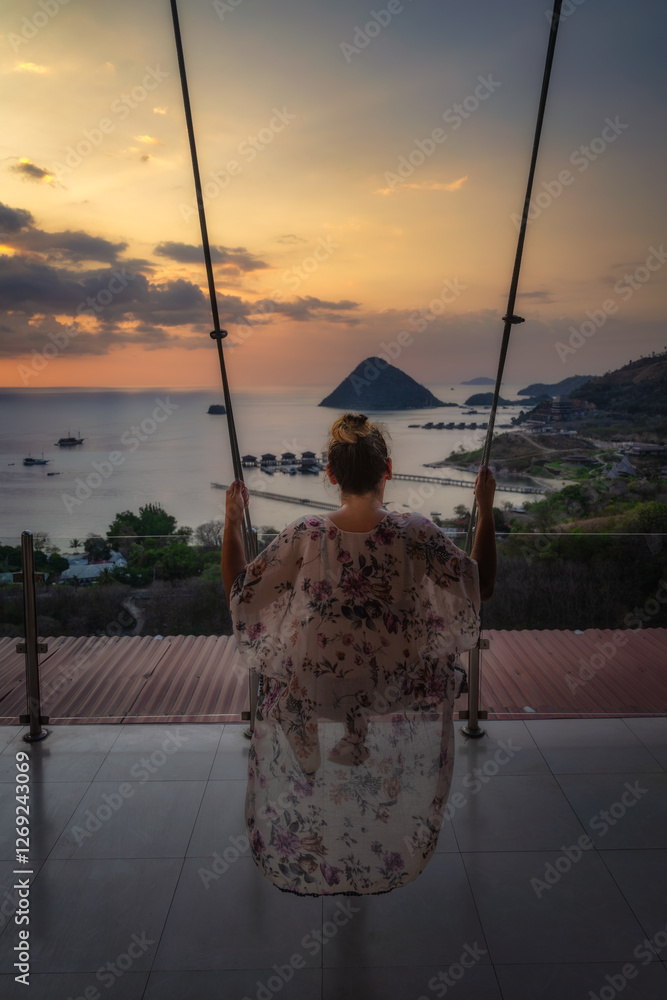A mesmerizing and beautiful sunset is artfully captured as a silhouetted figure joyfully enjoys swinging, overlooking the calming and tranquil ocean along with islands, Komodo, Indonesia