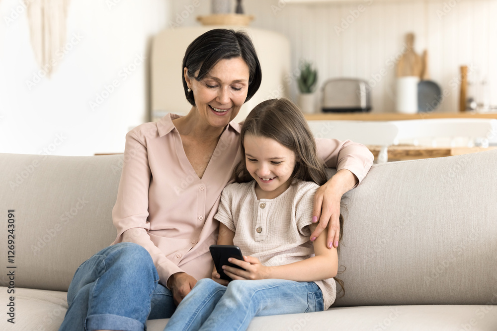 Digital tech and parental control. Cute little girl grandchild spend weekend at home sit on sofa with caring granny show funny blog on social media share photos on cell screen with engaged grandparent Digital tech and parental control. Cute little girl grandchild spend weekend at home sit on sofa with caring granny show funny blog on social media share photos on cell screen with engaged grandparent