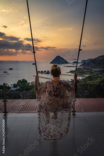 Obraz na płótnie A mesmerizing and beautiful sunset is artfully captured as a silhouetted figure joyfully enjoys swinging, overlooking the calming and tranquil ocean along with islands, Komodo, Indonesia w salonie