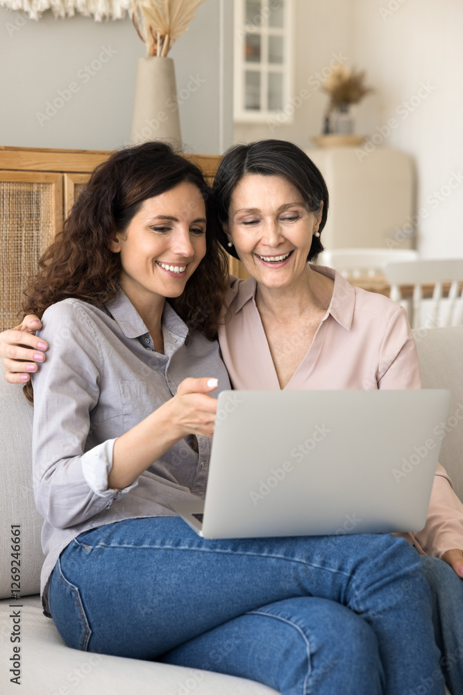Sharing digital experience. Smiling older age mother millennial daughter cuddle on sofa relax with notebook device watch funny video look through family photos make purchases at online ecommerce store Sharing digital experience. Smiling older age mother millennial daughter cuddle on sofa relax with notebook device watch funny video look through family photos make purchases at online ecommerce store
