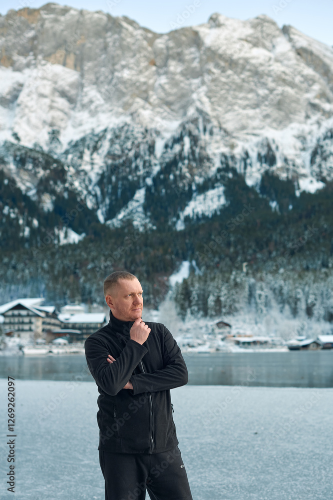 Fototapeta premium Contemplative Man in Black Outfit Admiring Snowy Alpine Landscape with Frozen Lake and Traditional Village. Serene Winter Mountain Scene Reflecting Tranquil Beauty of Nature in High Resolution