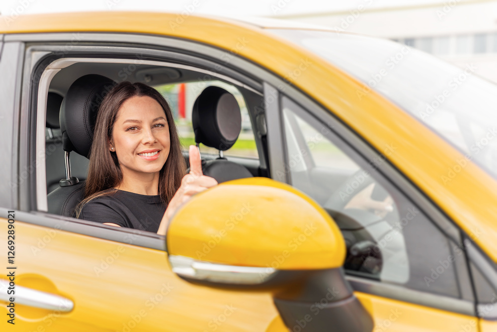 A young woman with long brown hair and a bright smile sits in the driver's seat of a yellow car, giving a thumbs-up gesture through the open window with a confident and positive expression.