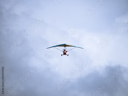 A small ultralight trike aircraft soaring in the sky with a pilot. The lightweight aviation design highlights recreational flying and personal air travel against a cloudy backdrop.