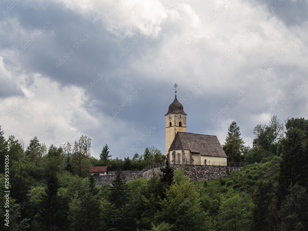 Fototapeta premium A historic church sits atop a lush green hill in Tarvisio, Italy, near the Alpe-Adria cycling route. The cloudy sky adds a dramatic touch to the serene and picturesque landscape.