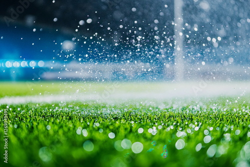 High-speed close-up of a sprinkler spraying water on a lush green soccer field at night, with glowing droplets creating a dramatic and atmospheric sports scene.

 photo