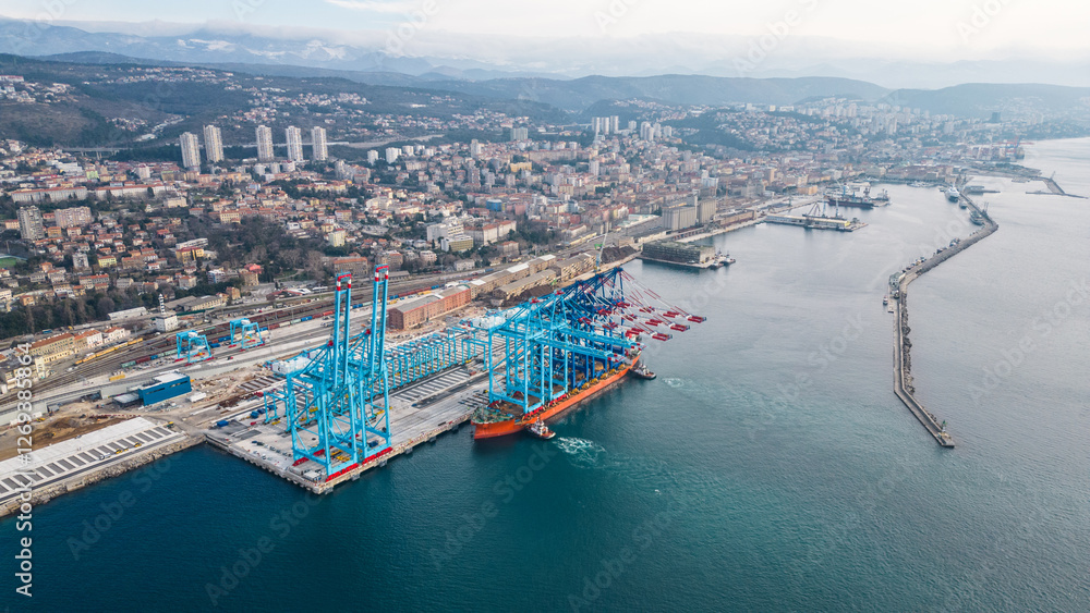 Naklejka premium An aerial drone view shows a cargo ship delivering new cranes to Rijeka’s modern cargo terminal port, boosting logistics and trade. This upgrade strengthens Rijeka’s role as a key Adriatic port