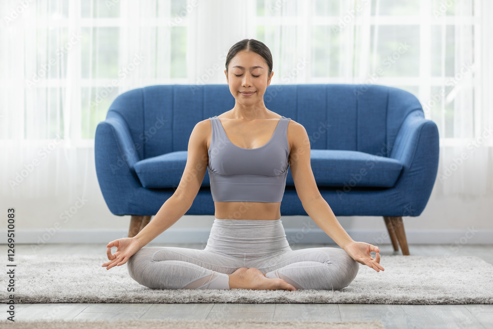 woman practicing yoga in serene living room, sitting cross legged on carpet, meditating with calm expression. background features blue sofa and bright natural light from large windows woman practicing yoga in serene living room, sitting cross legged on carpet, meditating with calm expression. background features blue sofa and bright natural light from large windows