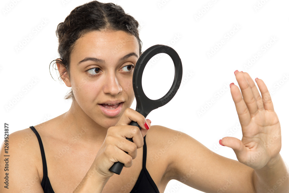 Young woman without makeup examining her nails with magnifying glass. Natural appearance, curly hair in bun. Curious expression. Studio portrait on white background Young woman without makeup examining her nails with magnifying glass. Natural appearance, curly hair in bun. Curious expression. Studio portrait on white background