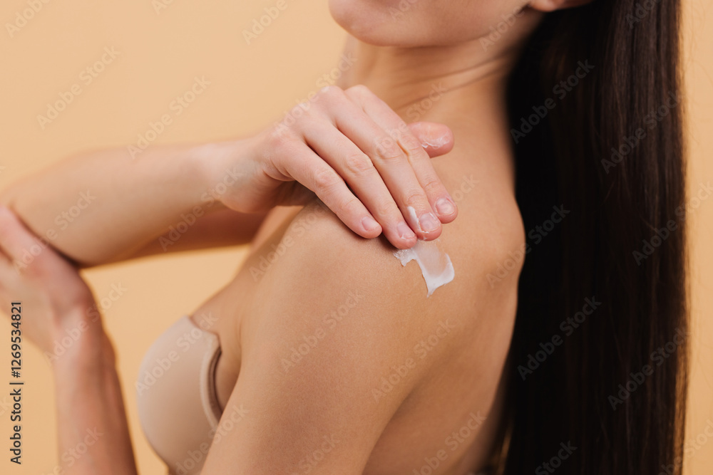 Fototapeta premium Woman applying moisturizing cream on her shoulder on a beige background.