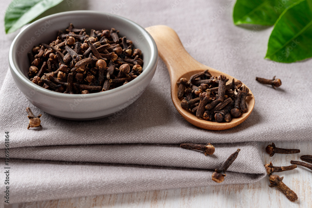 Bowl of organic clove buds close-up. Dry whole cloves spice in a small gray bowl and light wooden spoon over gray textile background. Spice and condiment concept. Bowl of organic clove buds close-up. Dry whole cloves spice in a small gray bowl and light wooden spoon over gray textile background. Spice and condiment concept.