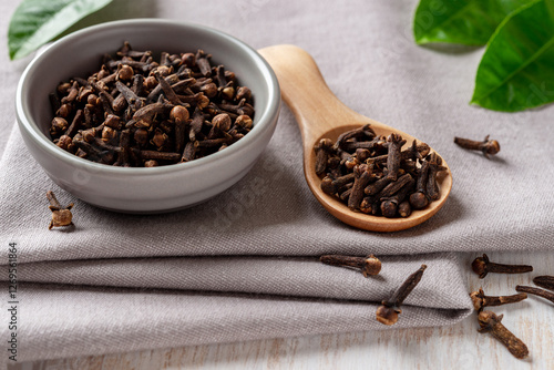 Bowl of organic clove buds close-up. Dry whole cloves spice in a small gray bowl and light wooden spoon over gray textile background. Spice and condiment concept.