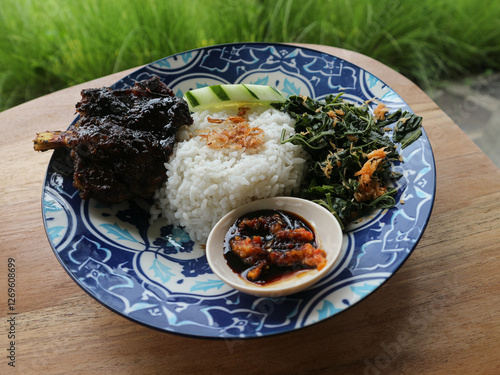 Grilled rib rice with sayur urap and soy sauce chili sauce served on a patterned blue plate with a green rice field background photo