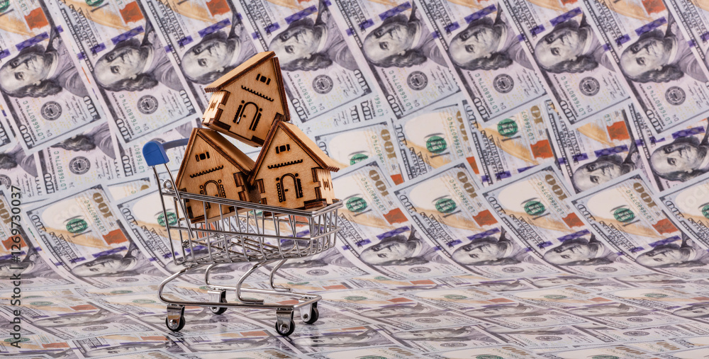 Naklejka premium Three symbolic wooden houses in a miniature supermarket shopping cart against a background of $100 bills