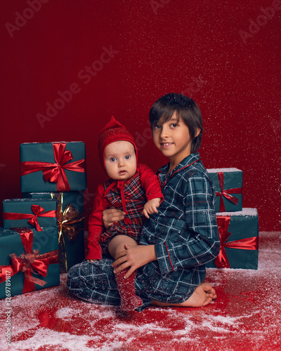 Obraz na płótnie brother and sister are sitting on the floor among green New Year gift boxes on a red background with snow. w salonie