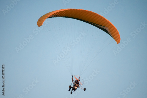 A paraglider soars through the sky above the mountains, carried by the wind. The paraglider is a bright orange color, and the pilot is silhouetted against the sky. The mountains are green and lush, an