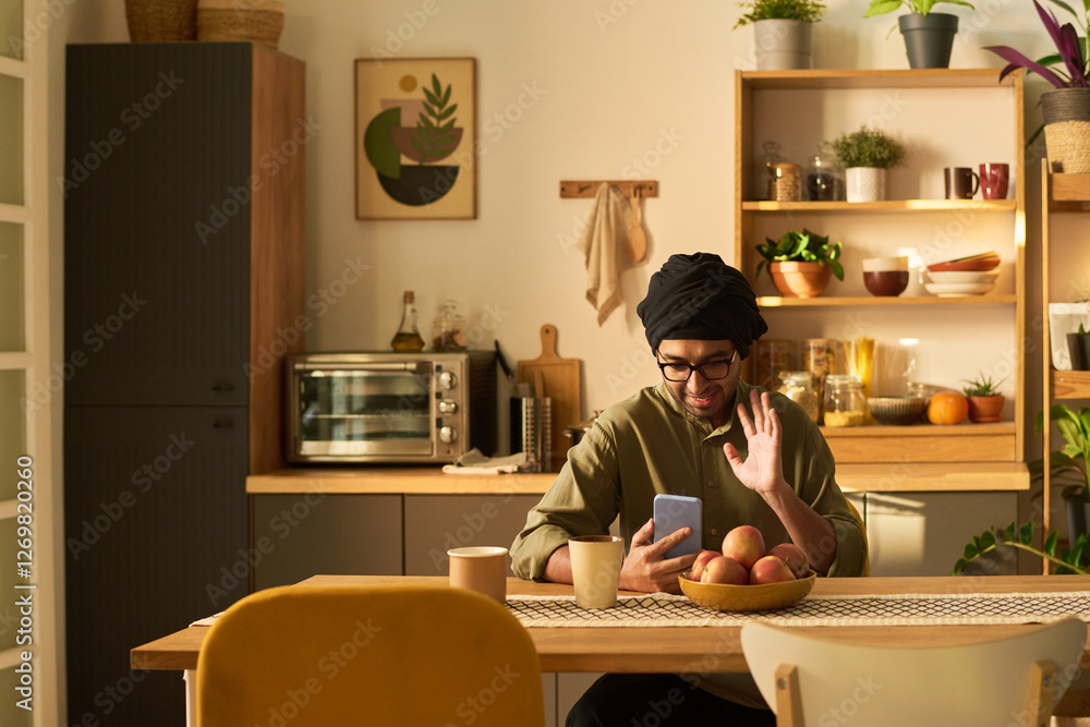 Man sitting in cozy kitchen, holding smartphone while touching beard and sitting at table with bowl of fruit. Warm ambient lighting highlighting kitchen decor and wooden elements Man sitting in cozy kitchen, holding smartphone while touching beard and sitting at table with bowl of fruit. Warm ambient lighting highlighting kitchen decor and wooden elements