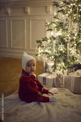 Obraz na płótnie One little girl in a red suit sits on a white carpet at the Christmas tree at home for 1 year. w salonie