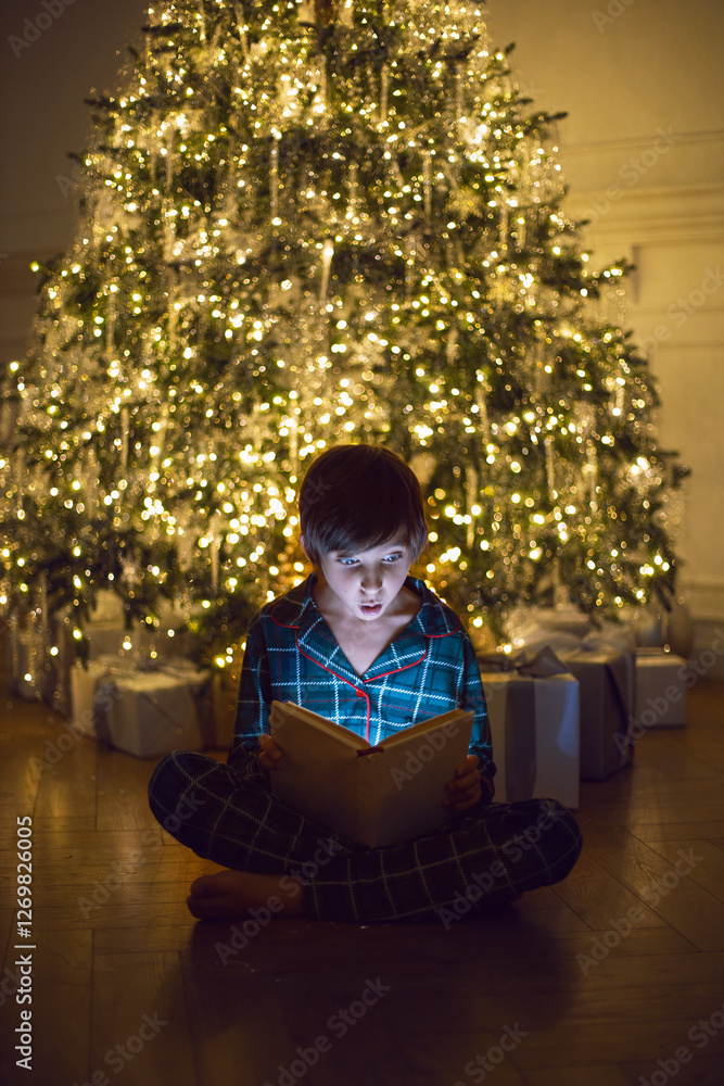 A happy 7 year old boy in green pajamas sit in the evening at a Christmas tree with garlands and Christmas gifts. A boy reads a magic book that glows