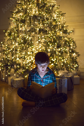 A happy 7 year old boy in green pajamas sit in the evening at a Christmas tree with garlands and Christmas gifts. A boy reads a magic book that glows