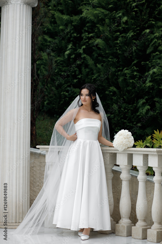woman in a wedding dress. She stands against the backdrop of green bushes. The dress is white, strapless, with a long skirt. She has a long veil on her head. woman in a wedding dress. She stands against the backdrop of green bushes. The dress is white, strapless, with a long skirt. She has a long veil on her head.