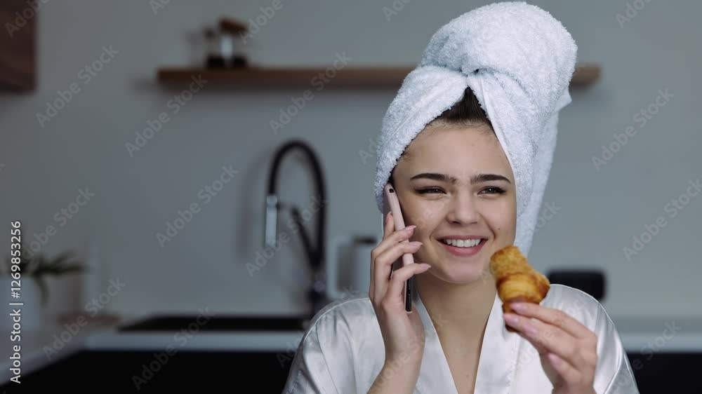 Young woman in kitchen during quarantine. Girl in bathing close after shower talking on phone and eating croissant. Young woman in kitchen during quarantine. Girl in bathing close after shower talking on phone and eating croissant.