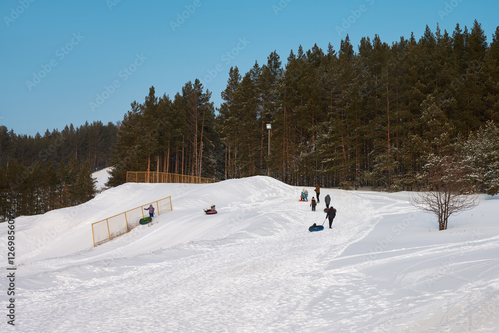 A tubing track at a ski resort. A group of people with children and inflatable sleds climbing a snowy slope with picturesque and pine trees. A tubing track at a ski resort. A group of people with children and inflatable sleds climbing a snowy slope with picturesque and pine trees.